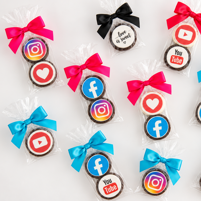 Twelve small round cookies decorated with colorful social media icons, individually wrapped in clear cellophane bags and tied with bright colored ribbons, arranged on a white background.