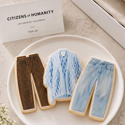 Three shaped sugar cookies styled to look like clothing pieces are arranged on a white plate: a dark brown textured pair of pants, a light blue knit cardigan, and light blue jeans. A white card reading "CITIZENS of HUMANITY LOS ANGELES, CALIFORNIA POP-UP" sits behind the plate on a light stone surface.