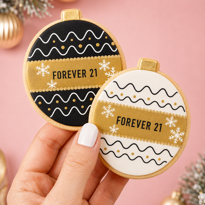 Two round Christmas ornament-shaped cookies held in a hand against a pink background with holiday decorations. One cookie is black with gold and white patterns and the text 'FOREVER 21', and the other is white with black and gold patterns and the same text.