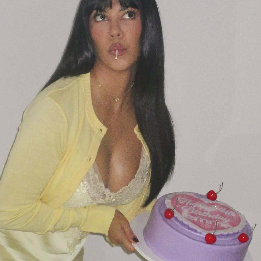 A woman with long black hair poses while holding a lavender birthday cake decorated with cherries on a white background.