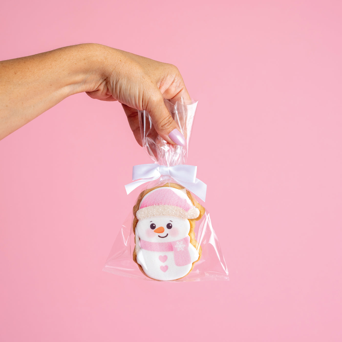 Hand holding a cookie shaped like a snowman in clear cellophane with a white ribbon against a pink background