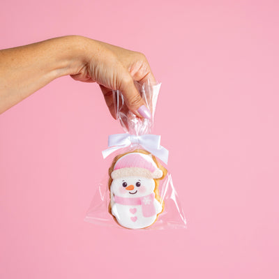 Hand holding a cookie shaped like a snowman in clear cellophane with a white ribbon against a pink background